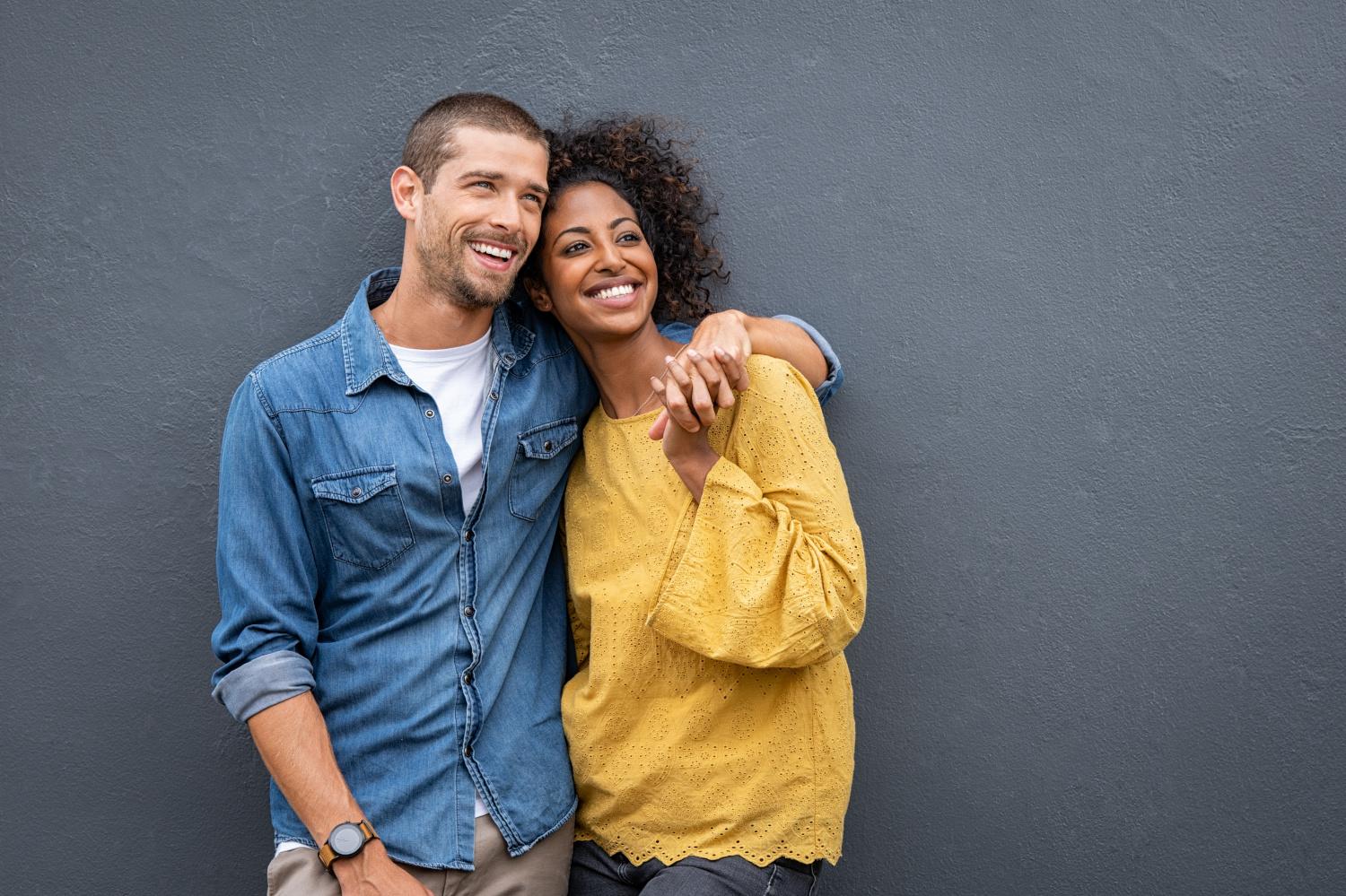Young multiethnic couple in love isolated on grey background looking up and thinking about their future together. Smiling man and african woman in casual hugging and looking away while planning the future. Cheerful couple holding hands while leaning on wa