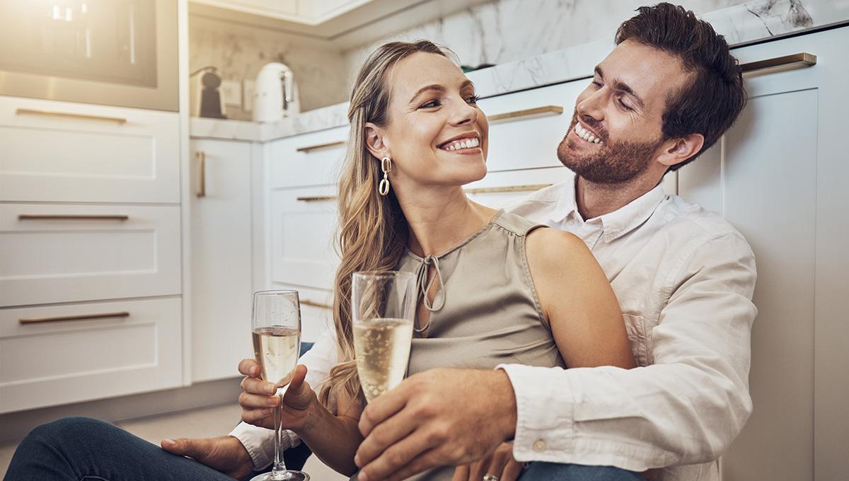 Celebrate, love and couple with champagne in kitchen for honeymoon, anniversary and valentines day. Happy, relax and young man and woman drinking alcohol on the floor with conversation in marriage