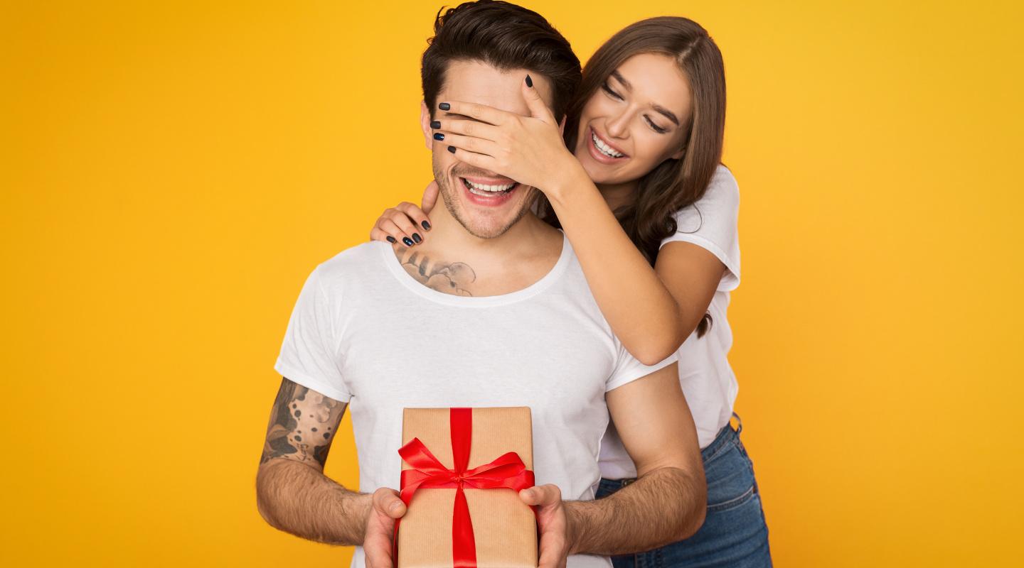 Girl covering her boyfriend eyes and giving him present on yellow background