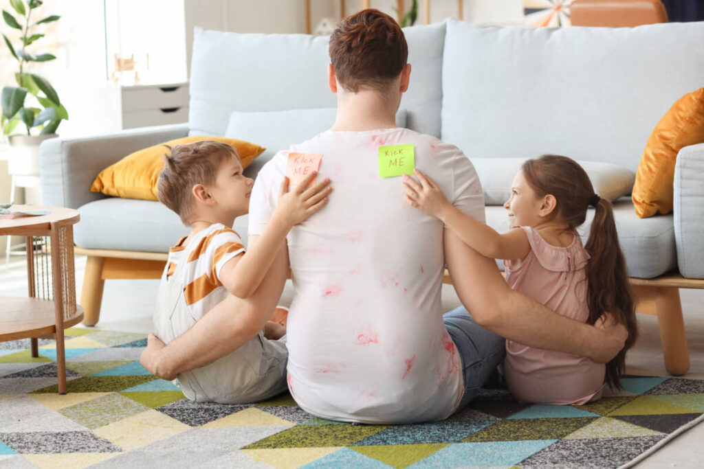 Little children sticking KICK ME stickers to their father's back at home. April Fool's Day prank